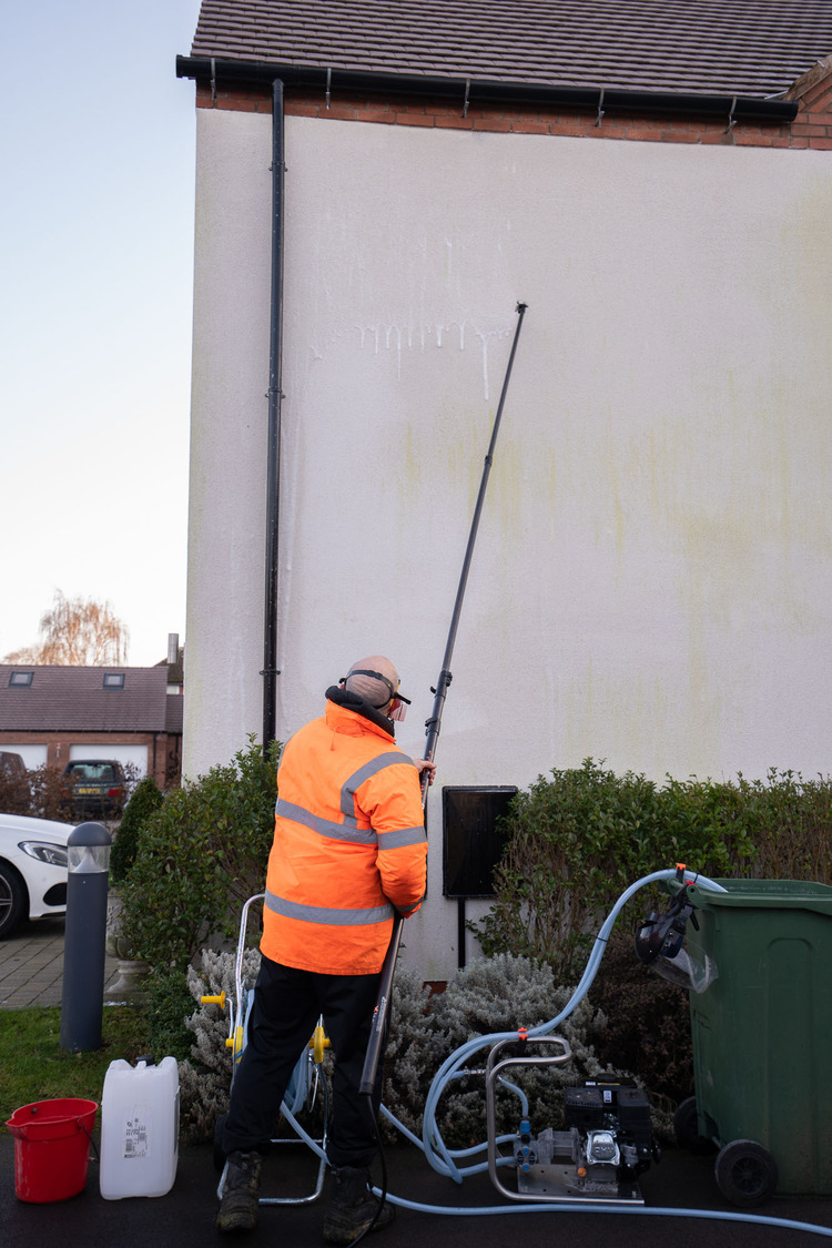 Render side gable mid-treatment with soft wash equipment in use, biocide being applied at low pressure to heavy algae staining